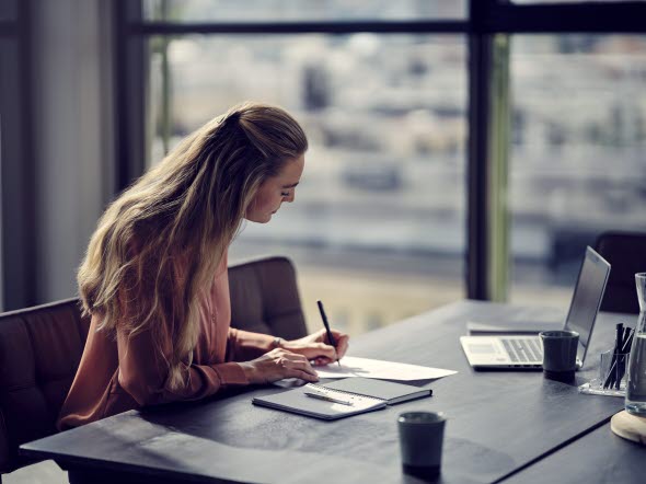 En kvinde sidder og skriver på et stykke papir ved et skrivebord En kvinde sidder og skriver på et stykke papir ved et skrivebord