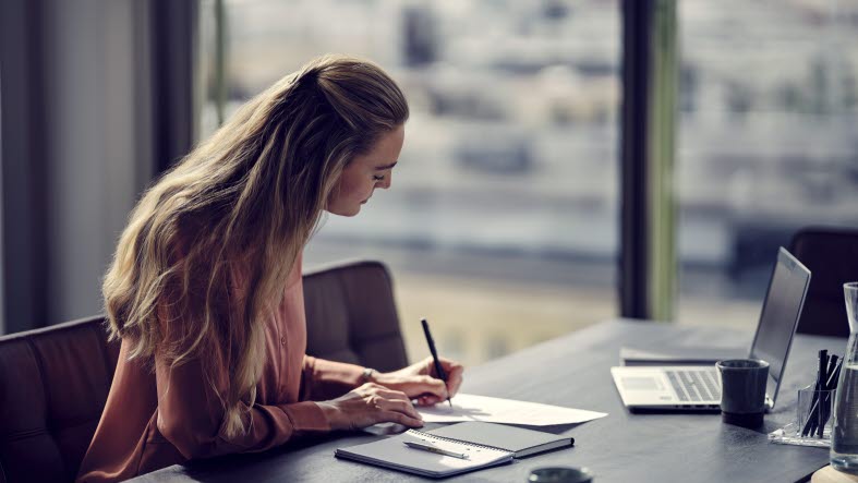 En kvinde sidder og skriver på et stykke papir ved et skrivebord