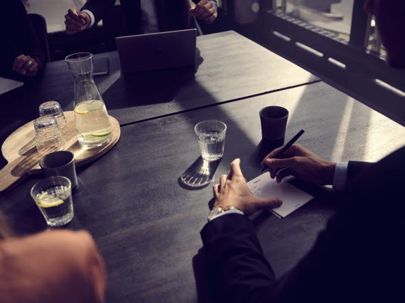 Mødedeltagere samlet om et bord, hvor man kun ser bordet og deres arme og hænder. En sidder med en laptop, en anden tager noter på en blok